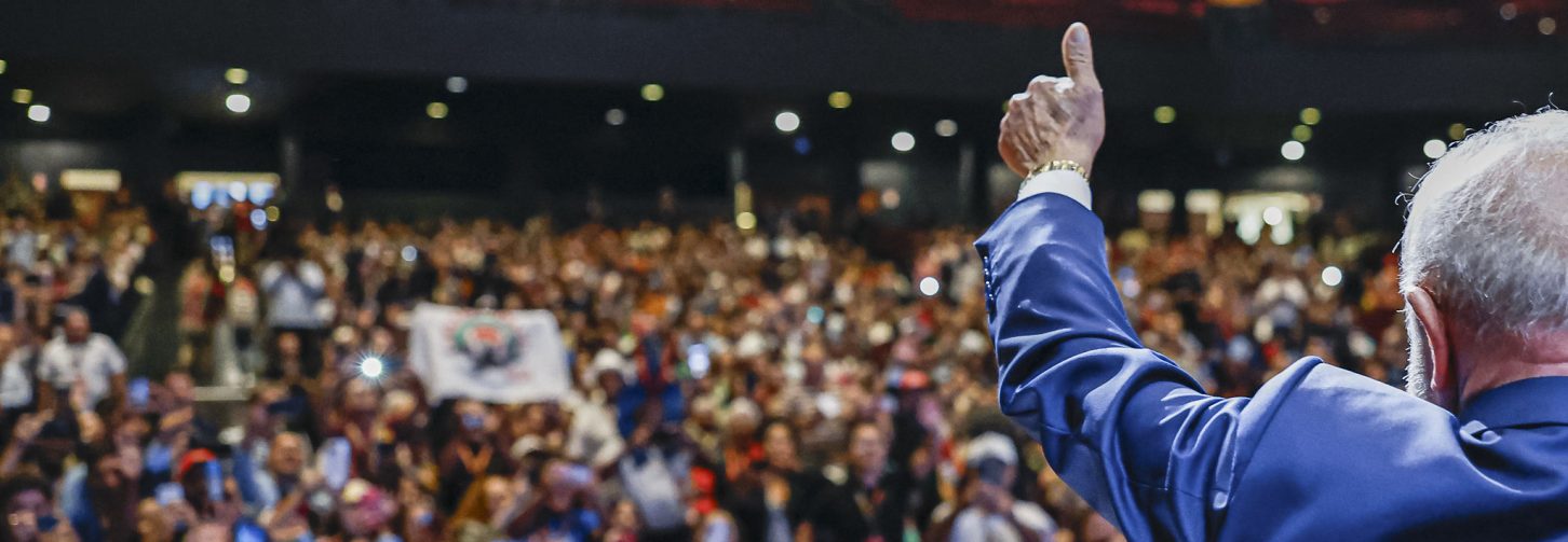 Em primeiro plano, visto de costas, Lula ergue o braço direito e acena para uma multidão. Ele veste paletó azul. Ao fundo, um grande público ocupa o espaço, com celulares levantados e bandeiras. O ambiente é amplo, iluminado por diversos pontos de luz no teto.