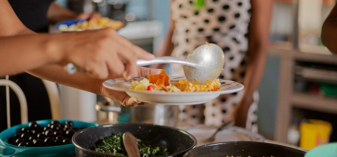 Uma pessoa se serve em uma cozinha com diferentes alimentos dispostos em travessas e tigelas. Ela segura um prato com arroz, salada e outros acompanhamentos enquanto usa uma colher para completar a refeição. Ao fundo, outra pessoa aguarda.