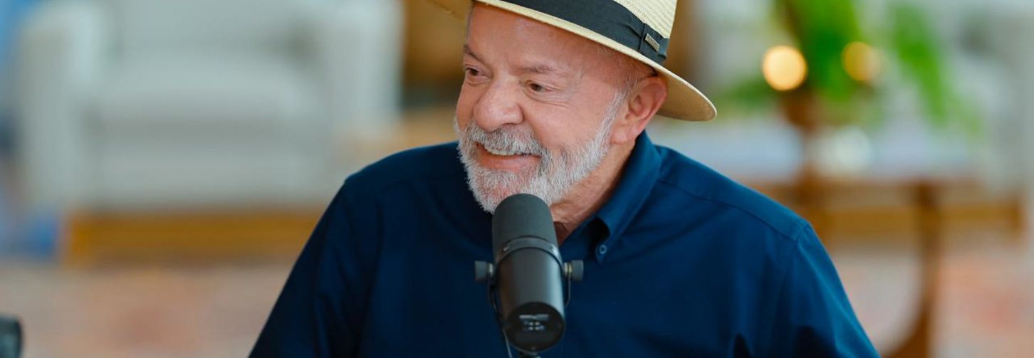 Presidente Lula com blusa azul escura, de chapéu e com ar sorridente falando ao microfone durante entrevista.