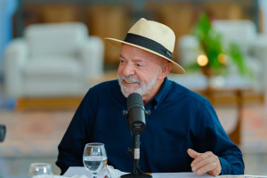 Presidente Lula com blusa azul escura, de chapéu e com ar sorridente falando ao microfone durante entrevista.