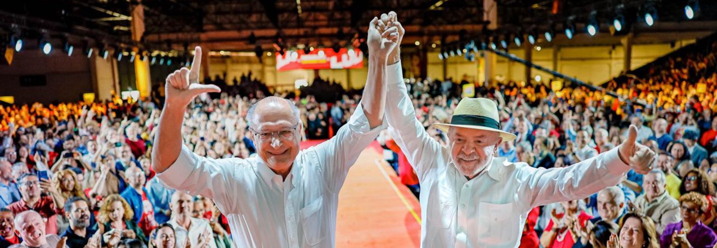Lula e Alckmin de mão levantadas em palanque com público ao fundo