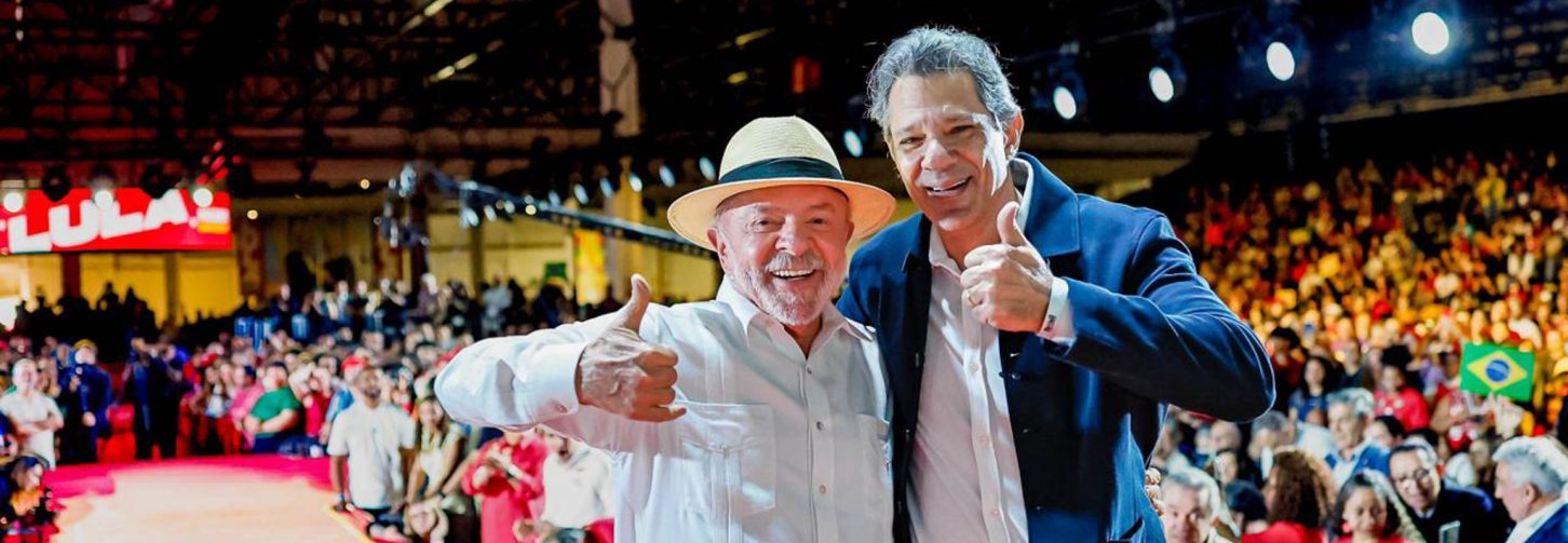 Presidente Lula, de camisa branca e chapéu Panamá, abraça candidato à governador de São Paulo, que veste terno azul. Os dois estão em palanque com tapete vermelho.