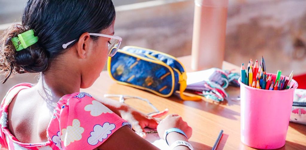 Uma estudante vista de costas escreve em um caderno sobre a carteira escolar. Ela usa óculos e uma blusa rosa estampada. Sobre a mesa há um estojo azul e amarelo, lápis de cor e outros materiais escolares.