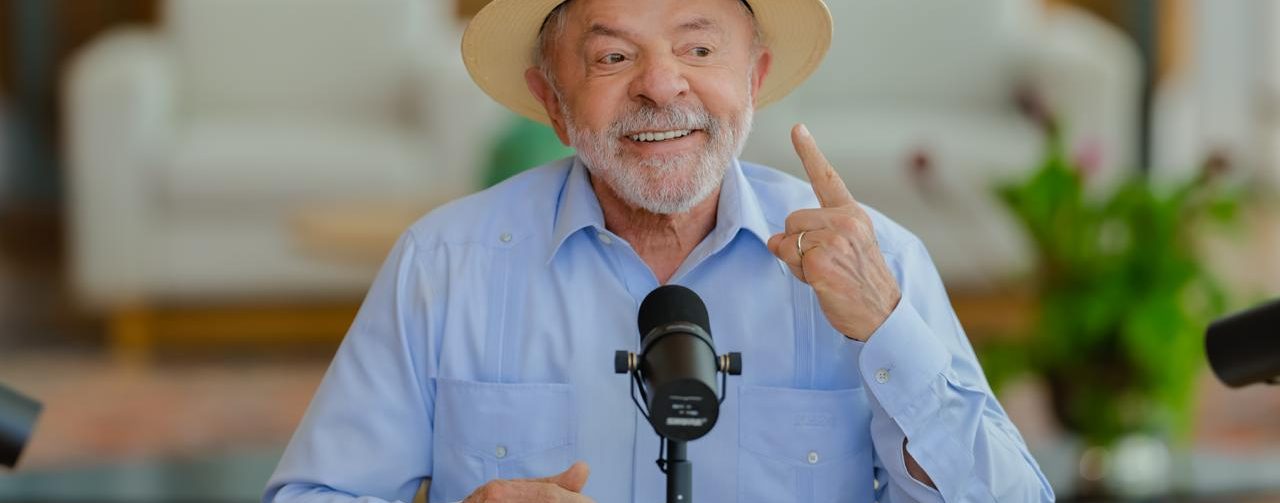Lula, sentado em frente à microfone, com camisa azul claro e chapéu Panamá