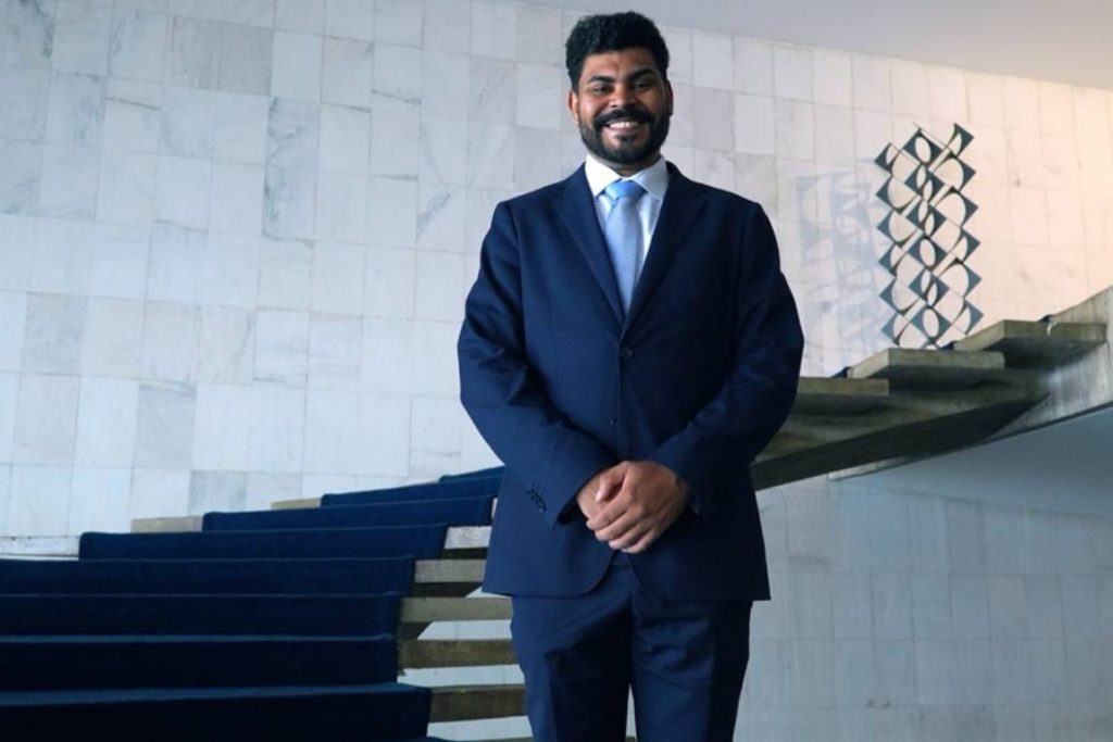 Um homem negro, de barba e cabelos curtos, sorri para a câmera. Ele veste terno azul-marinho, camisa branca e gravata azul-clara, e mantém as mãos unidas à frente do corpo. Ele está no Itamaraty. Ao fundo, há uma escadaria com carpete azul, paredes claras e uma escultura geométrica.