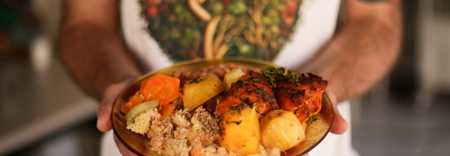Uma pessoa segura, diante do corpo, uma travessa redonda com uma refeição colorida, composta por arroz, carnes, legumes e verduras. Ela veste camiseta branca e avental com estampa de vegetais. Ao fundo, aparece o interior desfocado de uma cozinha.