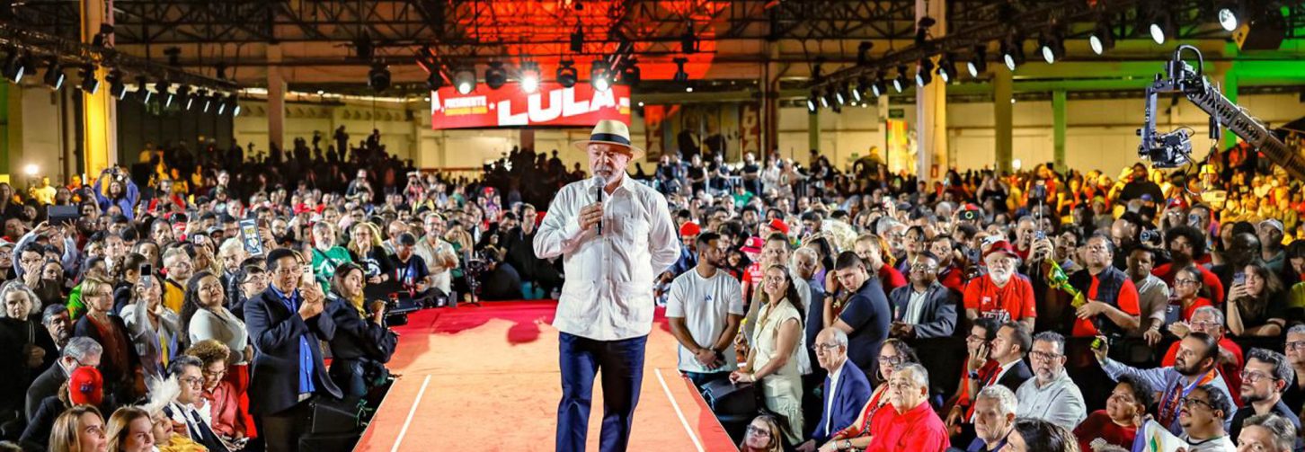 Lula, de chapéu e camisa branca, caminha por palanque vermelho discursando para plateia