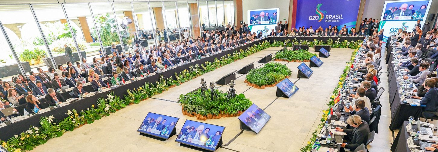 Uma grande mesa de reuniões durante o G20 realizado no Brasil, com o presidente Lula em primeiro plano nos telões do evento
