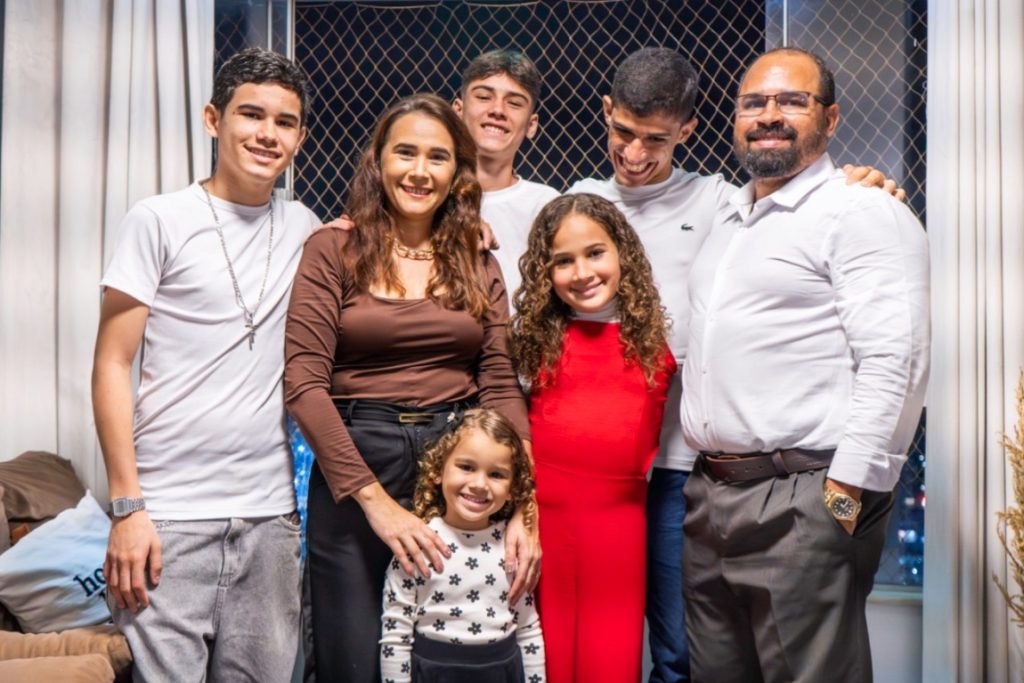 glicerio_Foto Mayara Vita Uma família de sete pessoas posa sorridente em uma sala. O grupo é formado por um homem e uma mulher adultos, três adolescentes e duas crianças. Eles estão abraçados e próximos uns dos outros. Ao fundo, há uma janela com cortinas claras e tela de proteção.
