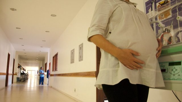 Gestante em corredor de hospital com enfermeira ao fundo. Ela segura a barriga e está de blusa branca e calça preta em primeiro plano