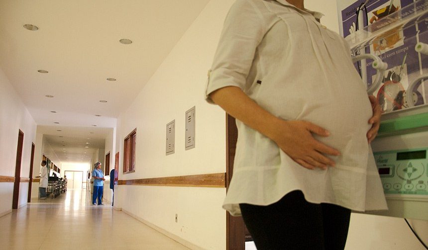 Gestante em corredor de hospital com enfermeira ao fundo. Ela segura a barriga e está de blusa branca e calça preta em primeiro plano
