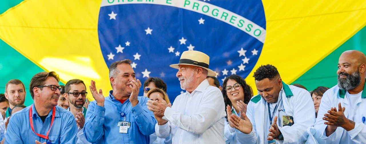 O presidente Lula à frente de especialistas na construção. do submarino nuclear brasileiro, com um painel grande ao fundo com a bandeira do Brasil projetada
