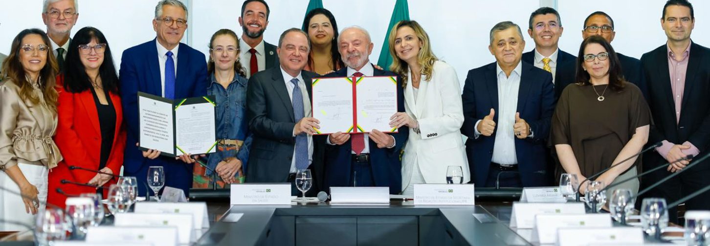 Várias pessoas atrás de uma mesa de reunião em pé. Lula ao centro com o papel da lei