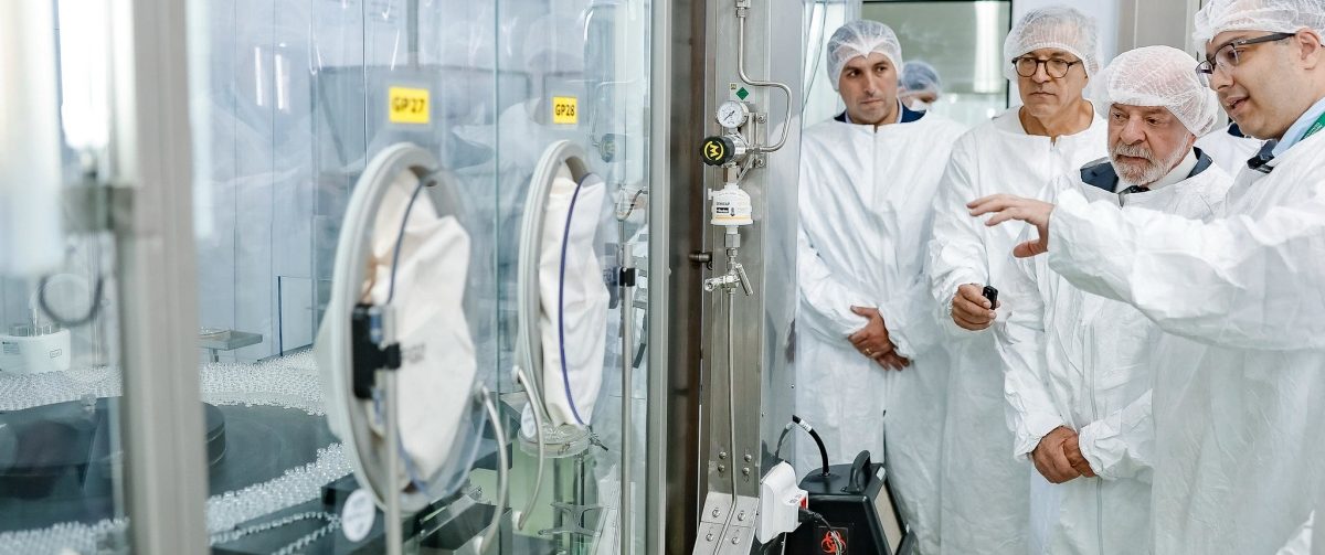 Imagem mostra o presidente Lula paramentado com avental de área de saúde, ao lado de técnico que mostra ao presidente a produção de vacinas no Instituto Butantan.